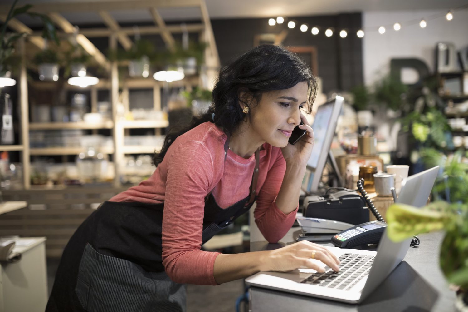 A shop owner on the phone while working at a laptop behind her counter
